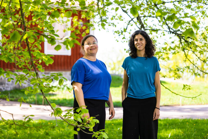 Assumed female interviewees Sandgren and Mazzi stand in front of a red wooden house, behind them luscious leafy tree.