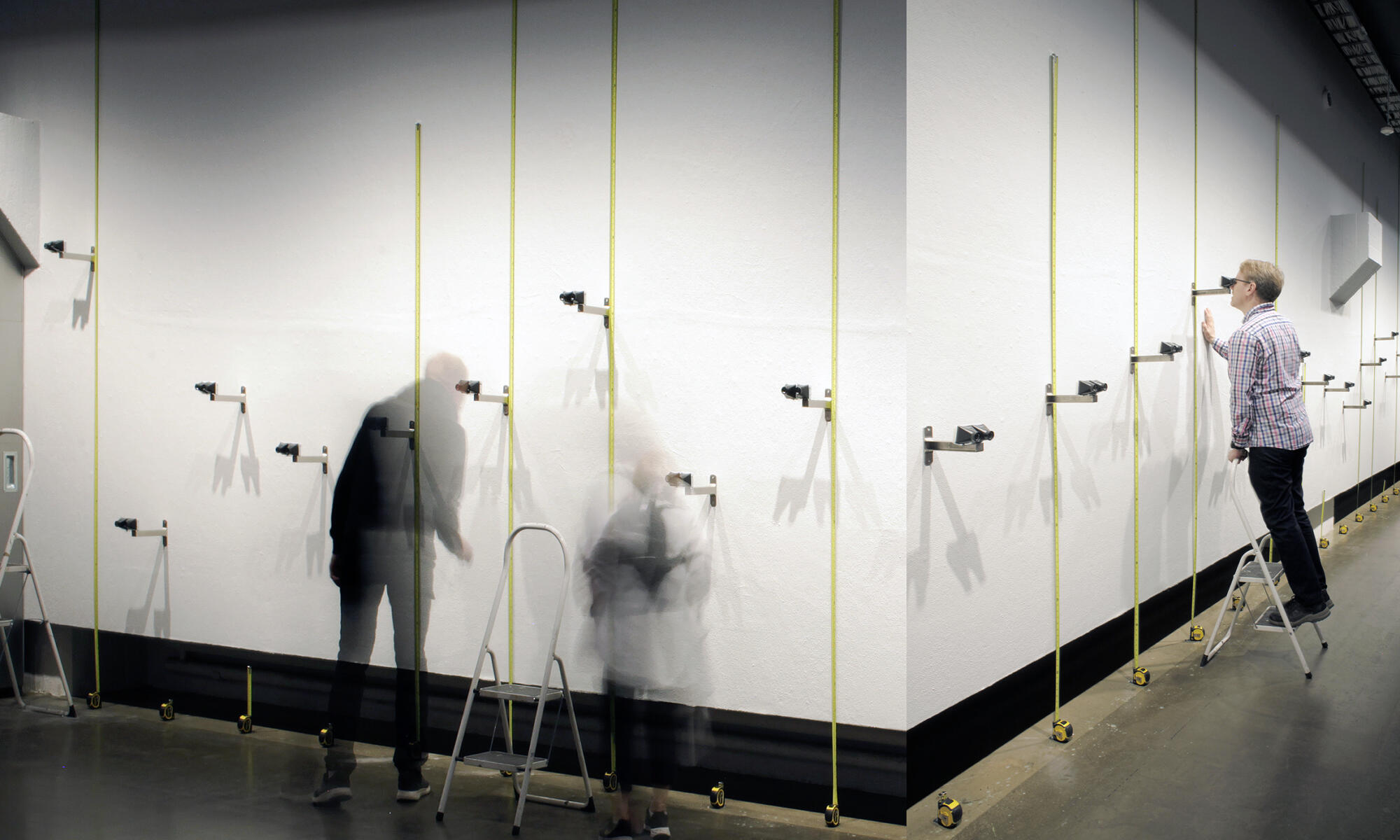 Pitkällä valotusajalla kuvattuja ihmishahmoja kuvattuna näyttelytilassa, jonka seinillä on kiikareita eri korkeuksilla.