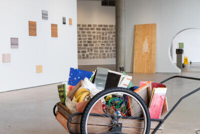 Exhibition view of a gallery space with a handcart full of various objects in the front.