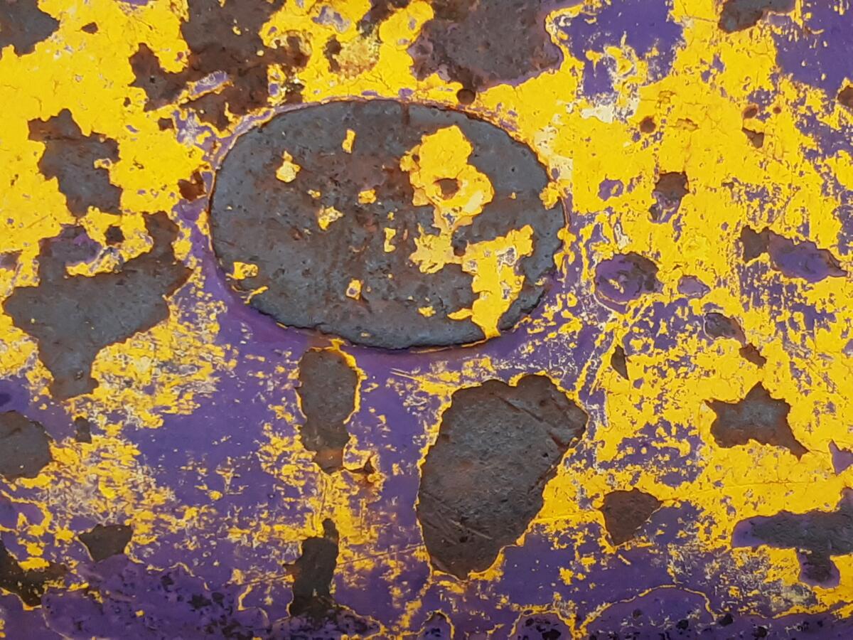 Ruosteinen, maalattu metallipinta, jossa voimakkaan keltaista maalin rippeitä ja alla violetti pohja sekä tummanruskean ruosteisia läikkiä.