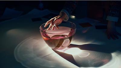 Käsi pitelee kiinni läpinäkyvässä maljan reunasta, jossa on vaalenapunaista nestettä. Valo taittuu maljasta heijastaen varjoja lattiaan.